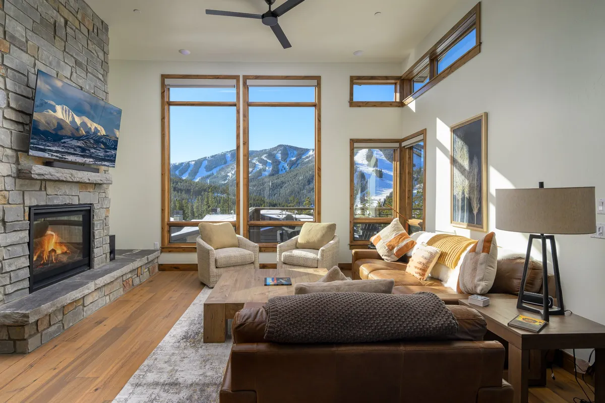 This image features a striking stone fireplace as the centerpiece of a comfortable seating area, with plush sofas and a wooden coffee table arranged for group gatherings. Large windows flood the space with natural light and offer sweeping views of the mountains, setting an inviting scene for cozy evenings or socializing. The room exudes warmth with its rich textures and spacious layout.