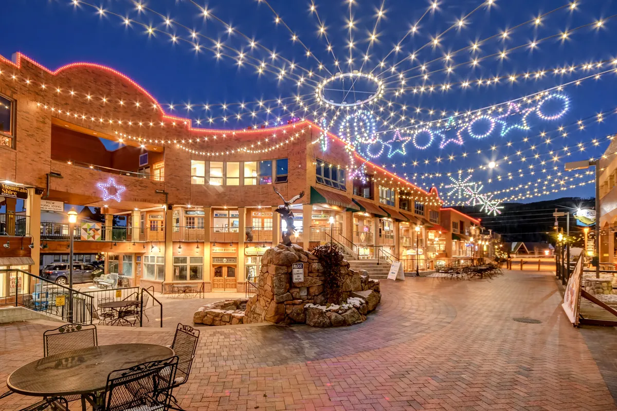 A lively outdoor plaza scene featuring string lights overhead, a stone fountain as a centerpiece, and buildings with large windows. People are seen strolling, dining, and socializing, while the outdoor seating areas invite guests to immerse themselves in the local atmosphere and community.