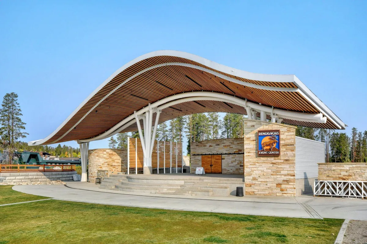 An exterior view of a striking curved roof structure with stone walls and wooden accents, set against a scenic backdrop. Event signage hints at upcoming community gatherings, outdoor concerts, or art exhibitions, inviting visitors to experience cultural and social events close to home.