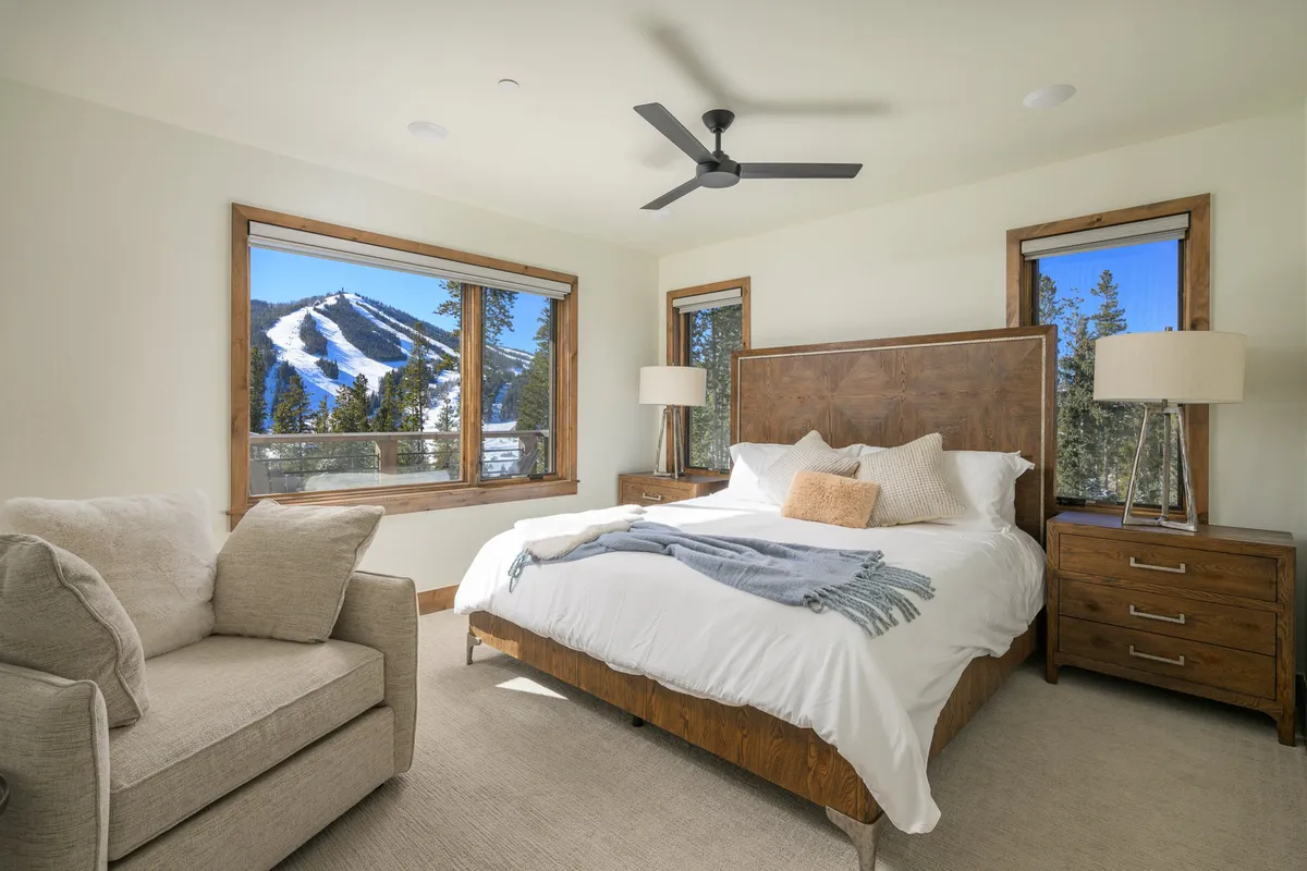 A tranquil bedroom featuring a large bed with crisp white bedding, a cozy armchair by the window, and expansive windows framing breathtaking mountain views. The decor is warm and inviting, accentuated by wooden furniture.