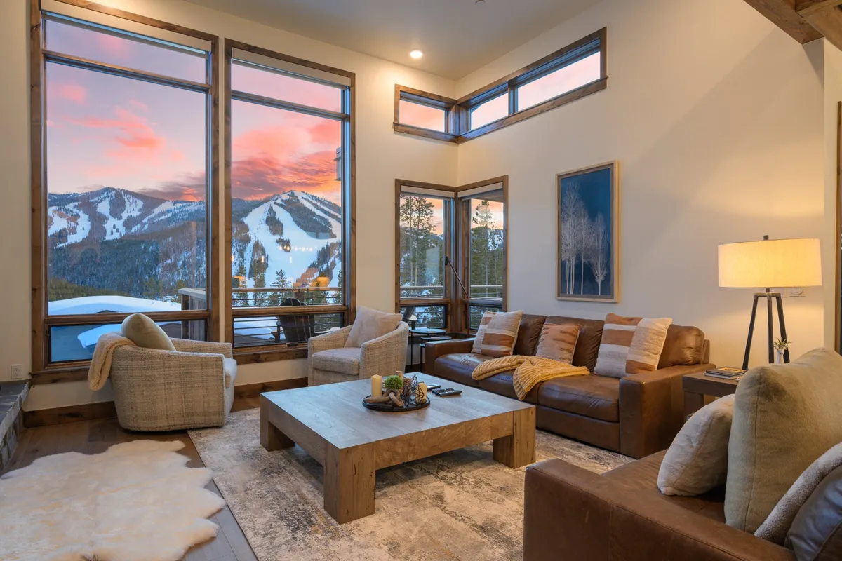 A plush seating area anchors the living room, with oversized windows framing breathtaking mountain vistas. The space is furnished for comfort and connection, complete with a wooden coffee table and artwork on the walls, creating a cozy yet sophisticated setting for relaxation or group activities.