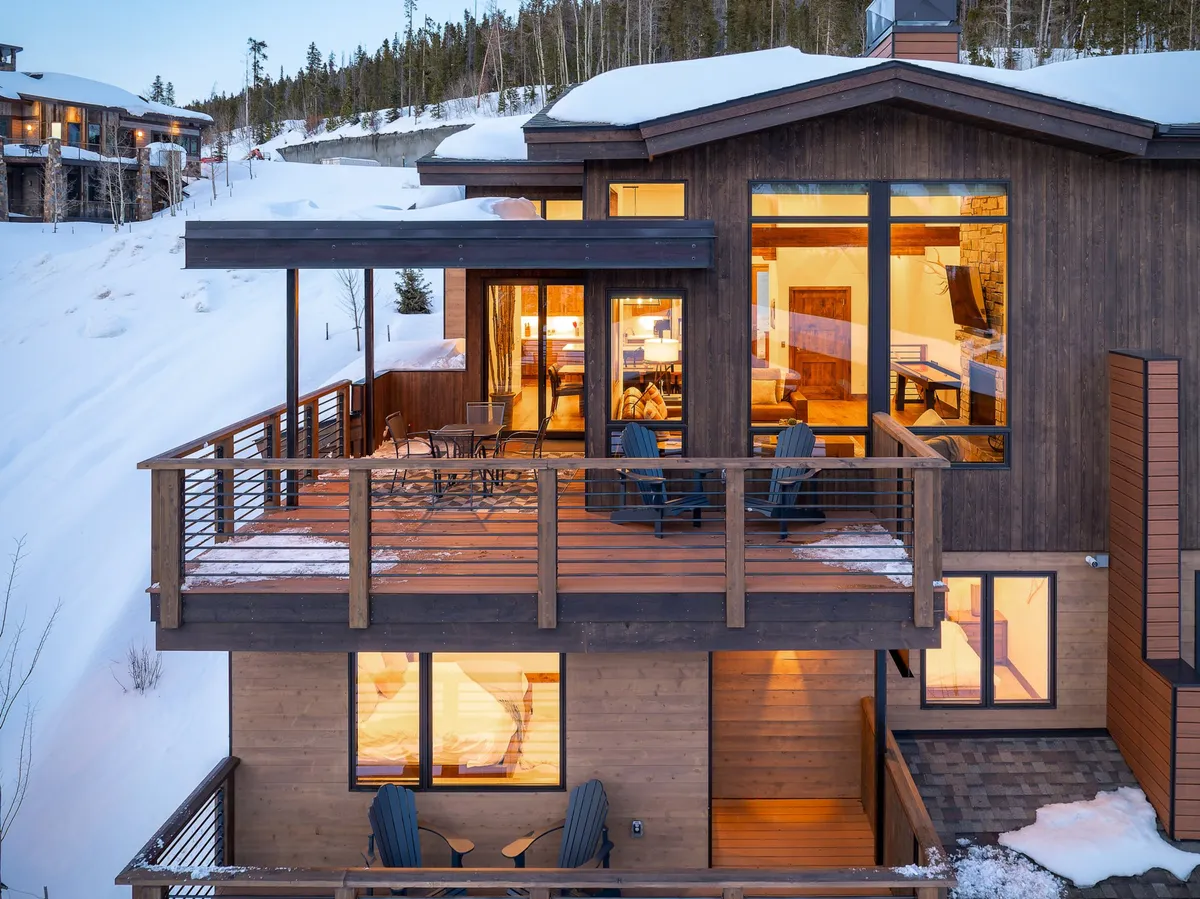 A wooden deck with outdoor seating overlooks snowy surroundings, with large windows connecting indoor and outdoor living. The deck is partially covered, inviting gatherings and scenic enjoyment throughout the year.
