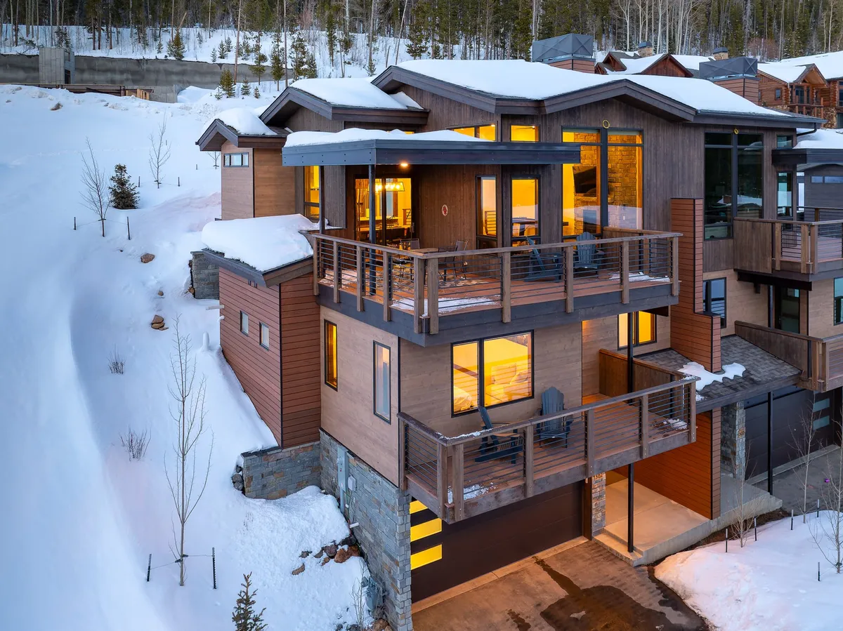 The image shows the inviting exterior of a luxury mountain villa nestled among snow-dusted trees, with a spacious wooden deck and expansive windows overlooking the snowy landscape. The scene evokes warmth and relaxation, perfect for watching falling snow or gathering outdoors with friends and family.
