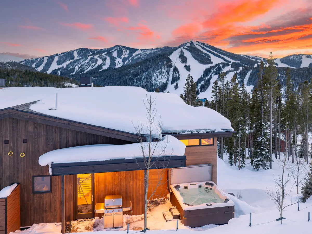 This exterior image features a hot tub set on a deck, partially covered by a snow-covered roof, with a dramatic mountain backdrop. The scene evokes relaxation after outdoor winter sports, star-gazing, and soaking up the serenity of the Rockies.