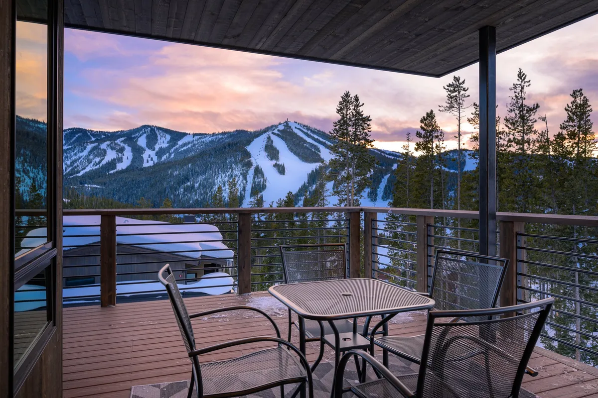 A peaceful view from a deck or patio, with comfortable outdoor furniture facing a sweeping mountain vista and tree-lined horizon. The golden-hour lighting suggests either sunrise or sunset, adding warmth and tranquility.