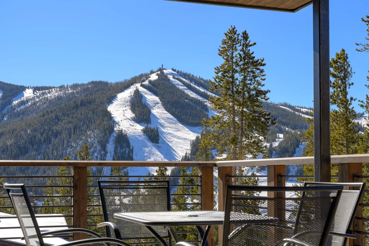 An inviting deck with comfortable seating overlooks snow-covered mountain slopes and pine trees. The scene captures both relaxation and adventure, with the ski slopes visible in the background, evoking the thrill of winter sports and the serenity of mountain living.