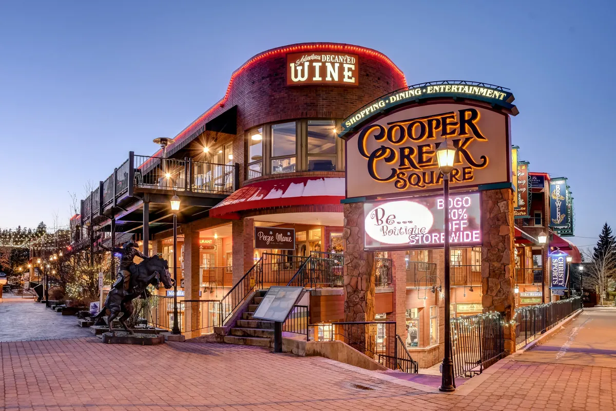 A lively exterior scene at Cooper Creek Square featuring a prominent sign, a wine store, outdoor stairway, and a horse sculpture. The area buzzes with activity, inviting visitors to shop, dine, and enjoy entertainment under the mountain sky.
