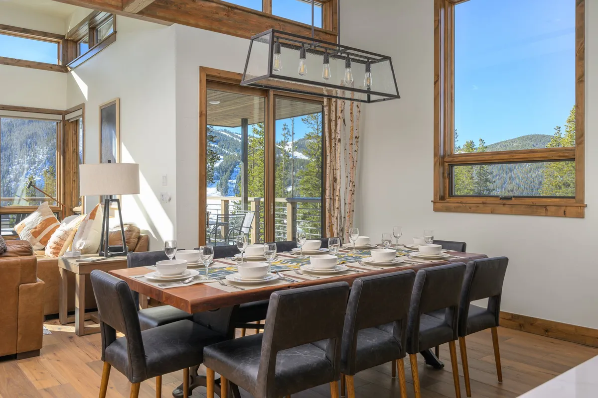 The dining area is shown with a large table set for a meal, surrounded by ample seating for a big group. Large windows reveal mountain views, while a nearby comfortable seating area suggests easy flow between dining and living spaces. The atmosphere is inviting and ideal for family gatherings or celebrations.